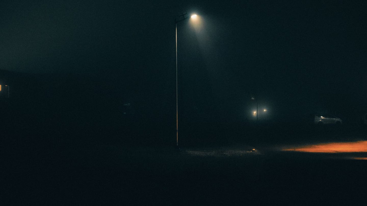 A dark city street at night, streetlights glowing against deep shadow